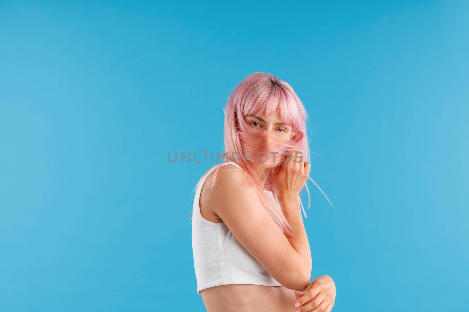 Attractive woman in white shirt looking at camera, hiding her face with pink hair while posing isolated over blue studio background by Yaroslav_astakhov