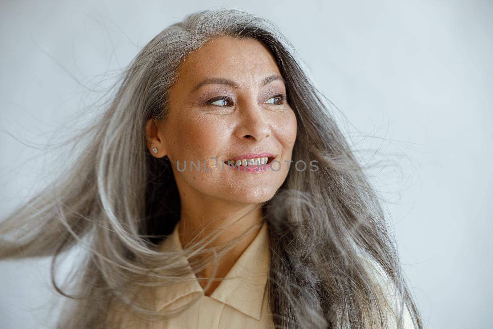 Happy middle aged Asian woman with flying grey locks stands on light background by Yaroslav_astakhov