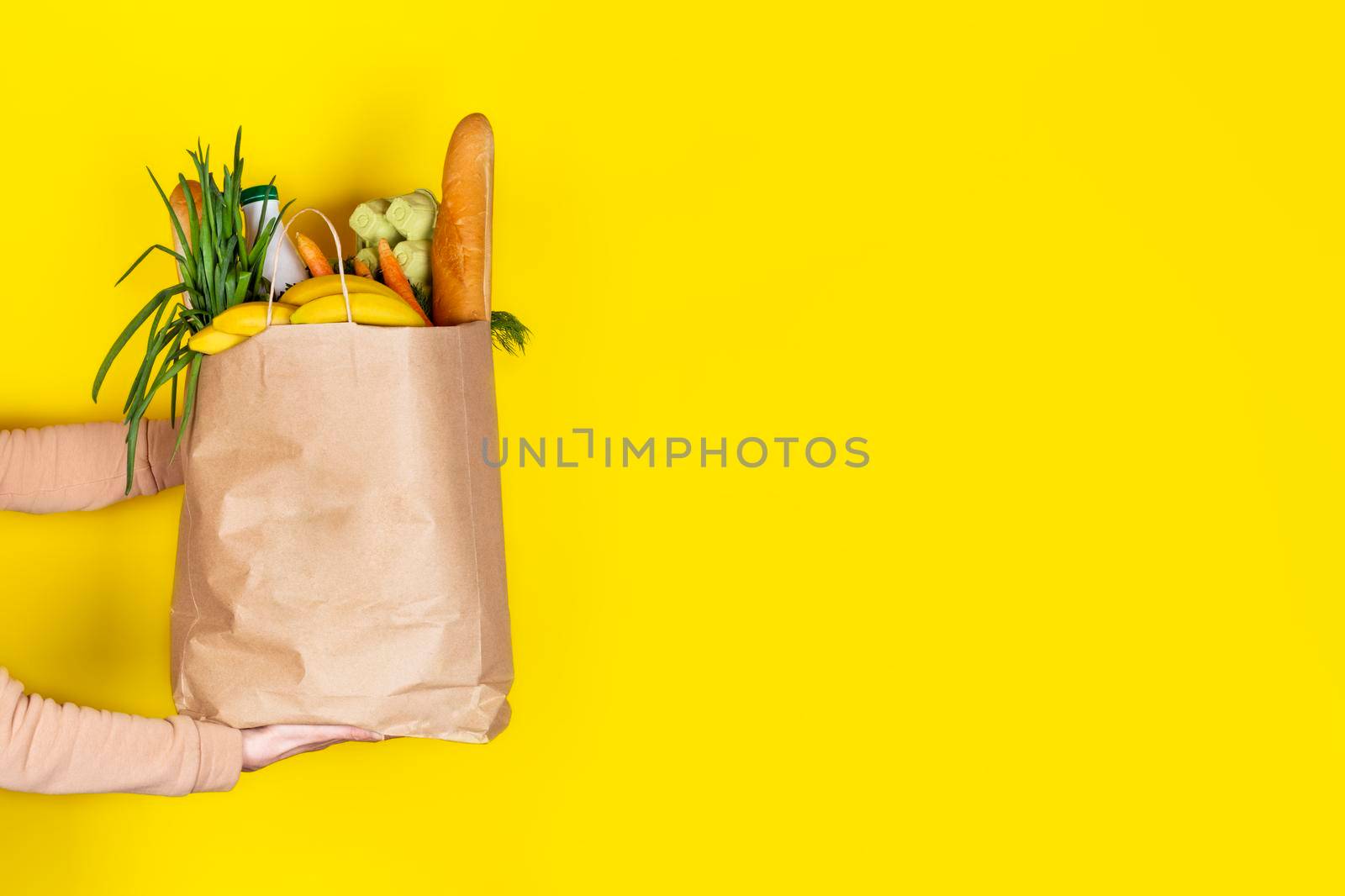 Food delivery or donation concept. Grocery store shoping. Girl or woman holds a paper bag filled with groceries such as fruits, vegetables, milk, yogurt, eggs isolated on yellow. Copy space