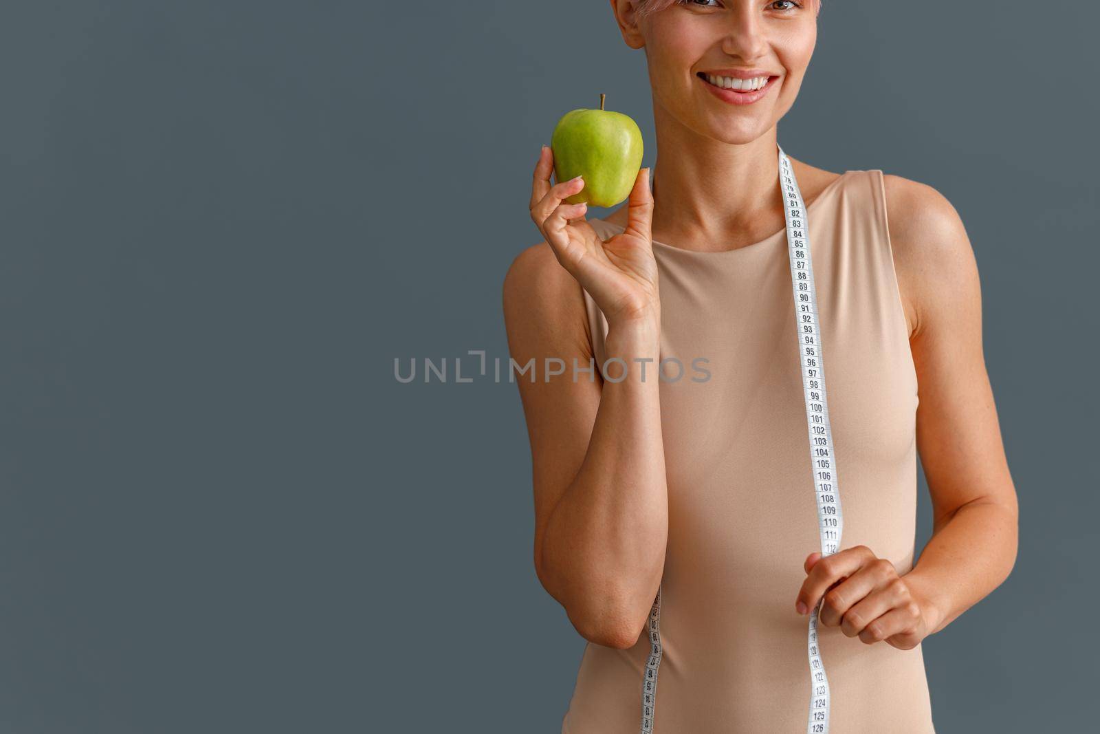 Cropped shot of young woman in beige bodysuit smiling at camera, holding green apple and measuring tape isolated over gray studio background by Yaroslav_astakhov