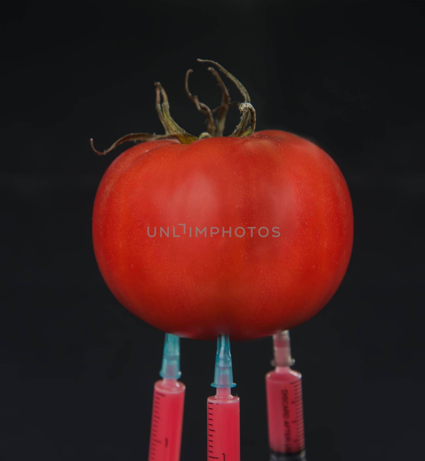 Red liquid in the syringe injected into tomato on a black background, close-up