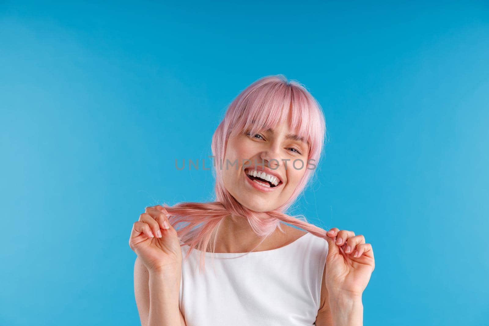 Happy female model playing with smooth straight pink hair, wrapping it around her neck while posing isolated over blue studio background. Beauty, hair care concept