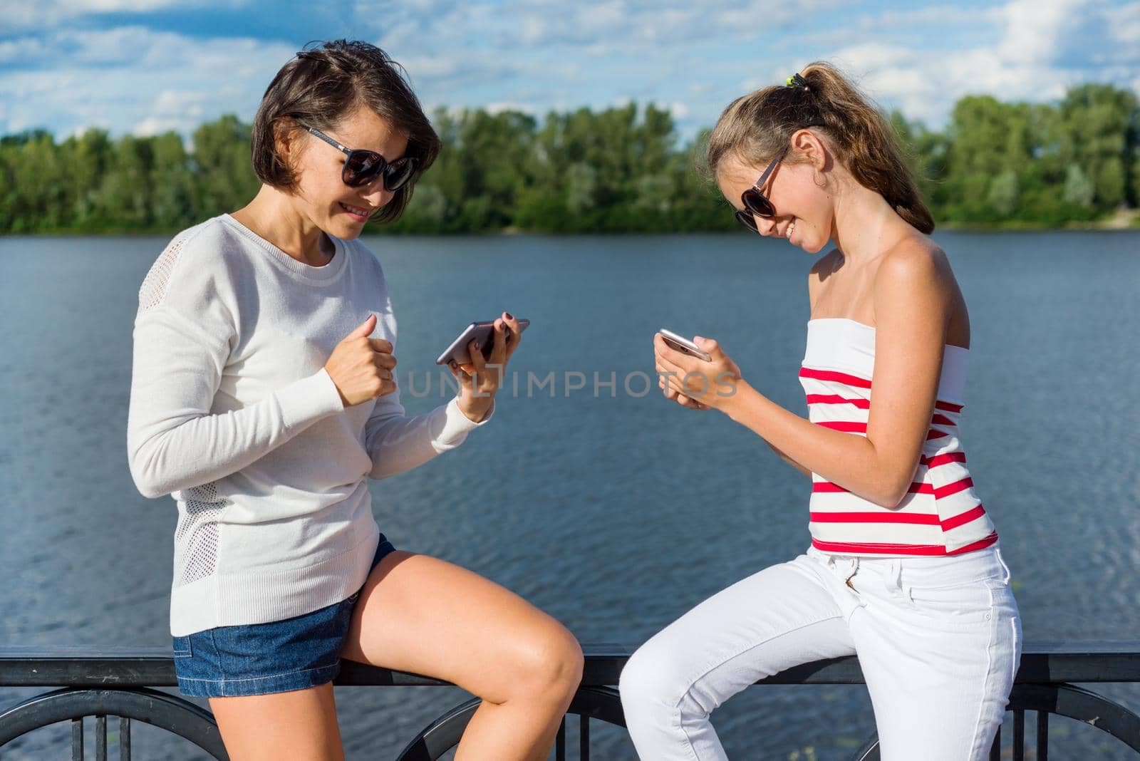 A pretty teenage girl and her beautiful mother use smartphones, view photos, talk and smile, on a sunny summer day in the city park