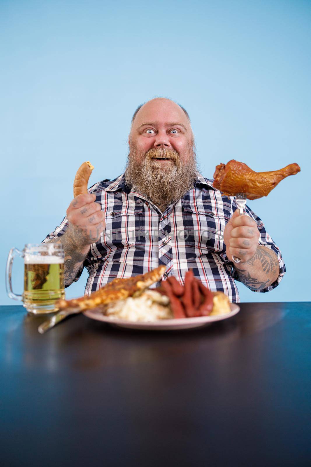 Cheerful bearded person with overweight holds smoked chicken leg and sausage at table with fat food and beer on light blue background in studio