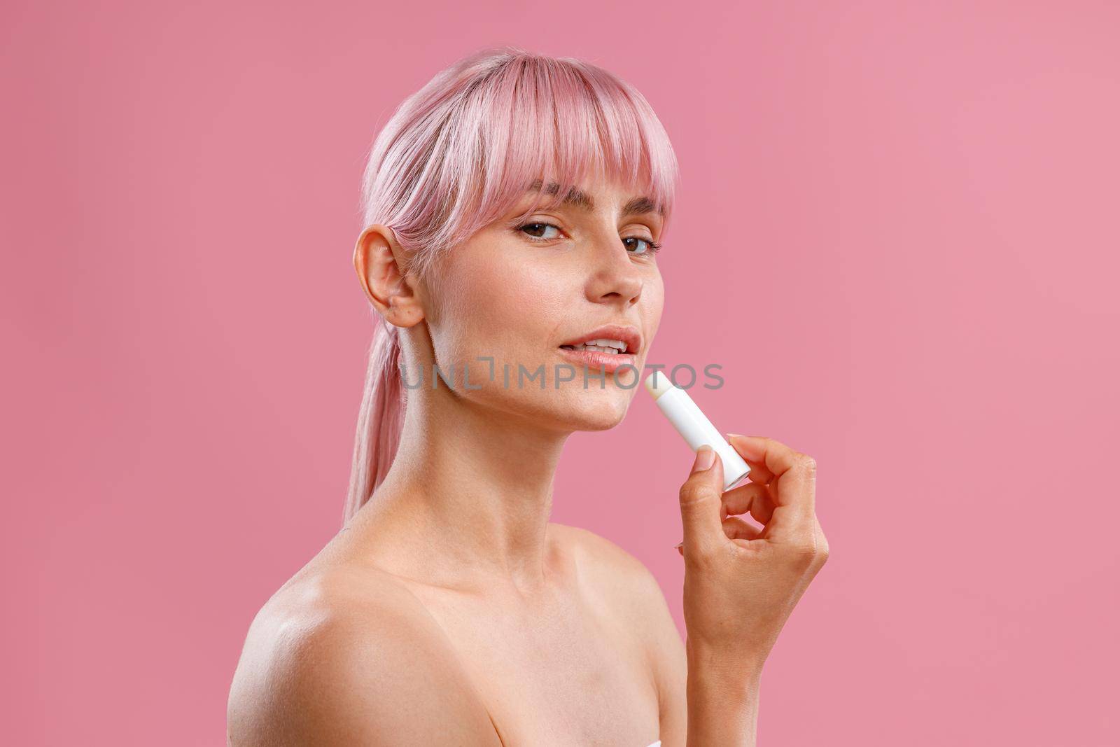 Portrait of beautiful young woman with naked shoulders holding lip balm near her lips isolated over pink background by Yaroslav_astakhov