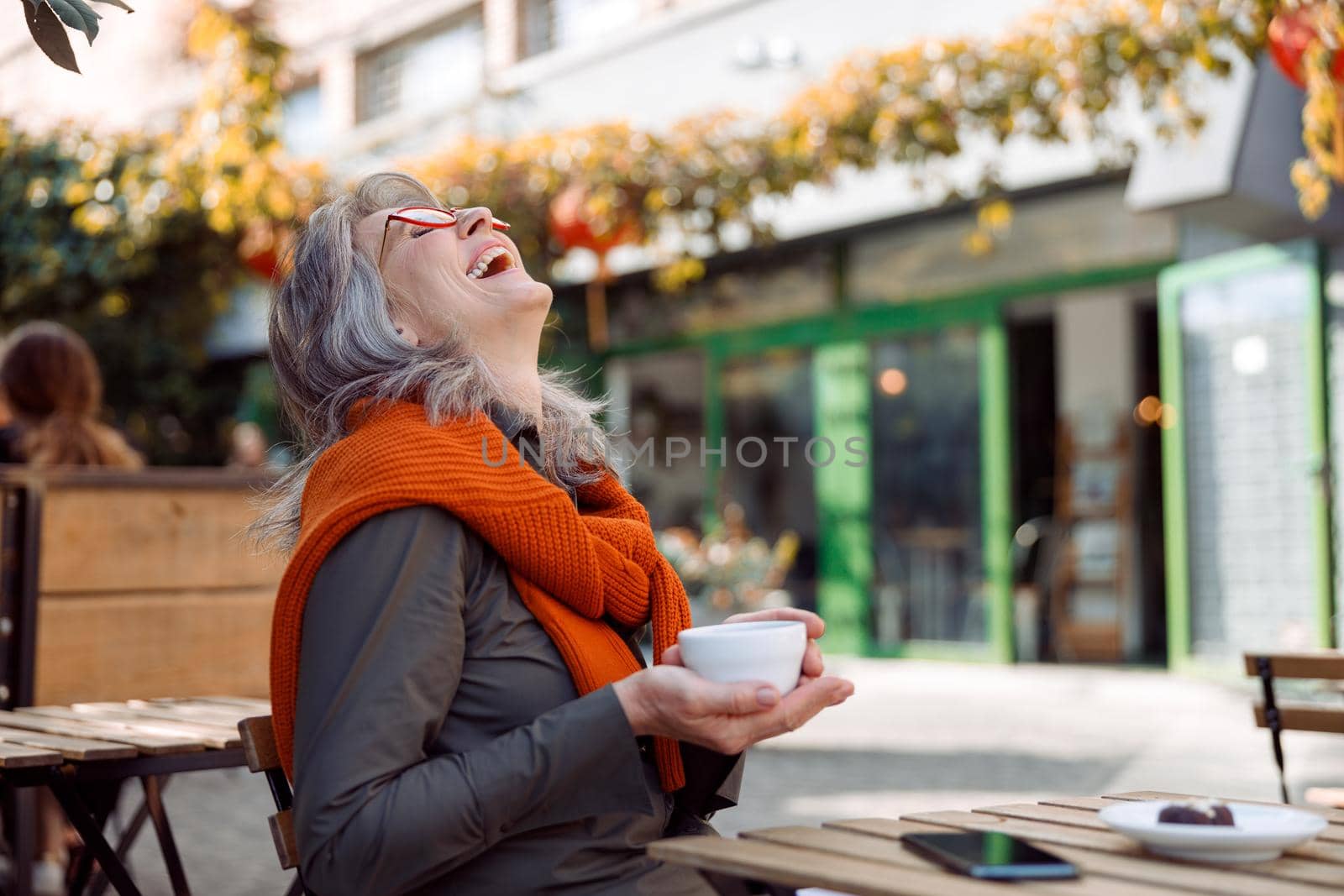 Cheerful senior lady with glasses and cup of tasty coffee laughs throwing back her head at table on outdoors cafe terrace on autumn day