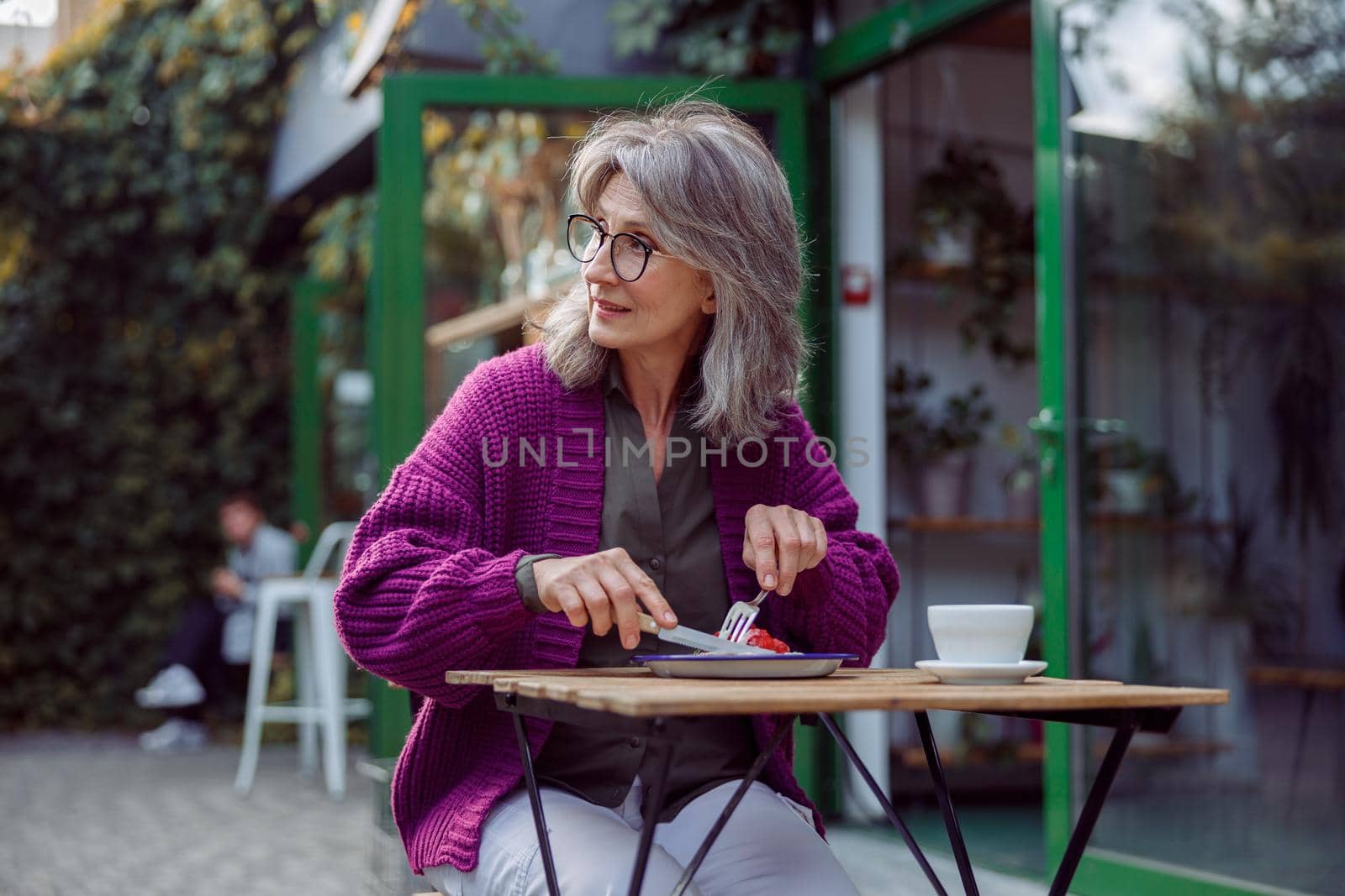 Silver haired mature woman in warm knitted jacket enjoys delicious dessert on outdoors cafe terrace by Yaroslav_astakhov