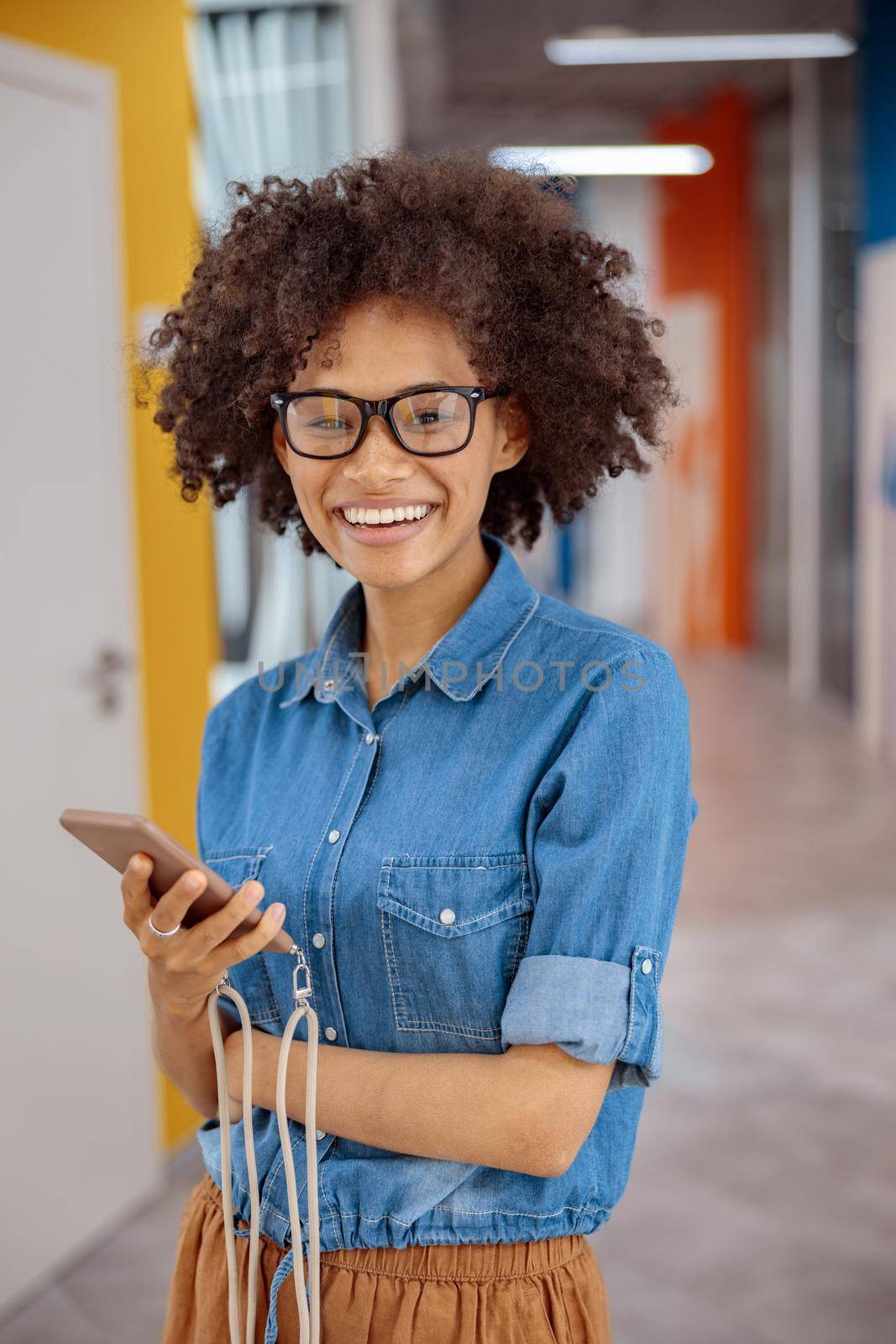 Afro American lady in glasses standing in the office and holding smartphone while smiling