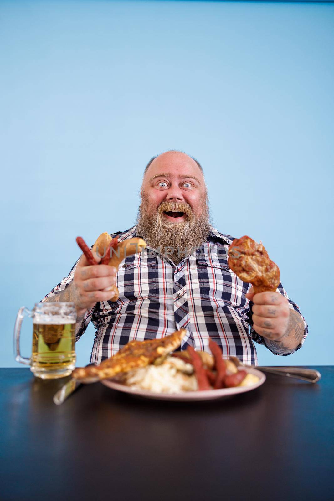 Excited obese man holds chicken leg and bunch of sausages sitting at table with greasy food and beer on light blue background in studio