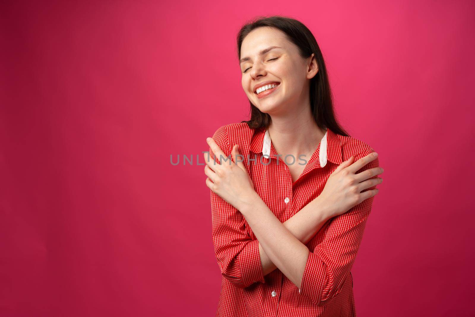 Joyful smiling young woman hugging herself against pink background, close up