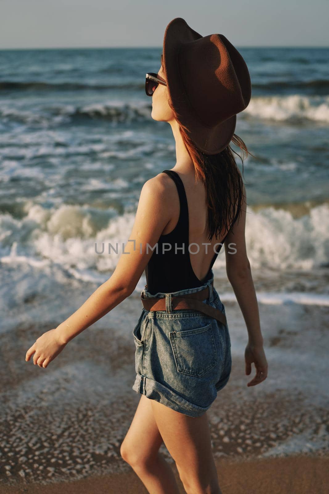 cheerful woman in sunglasses and a hat walks along the ocean by Vichizh