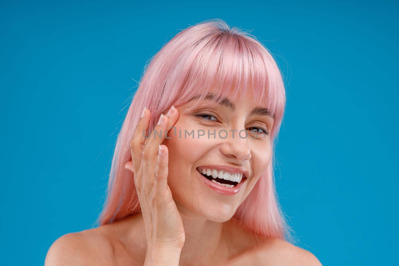 Portrait of smiling young woman with pink hair touching her perfect skin, looking at camera, posing isolated over blue studio background by Yaroslav_astakhov