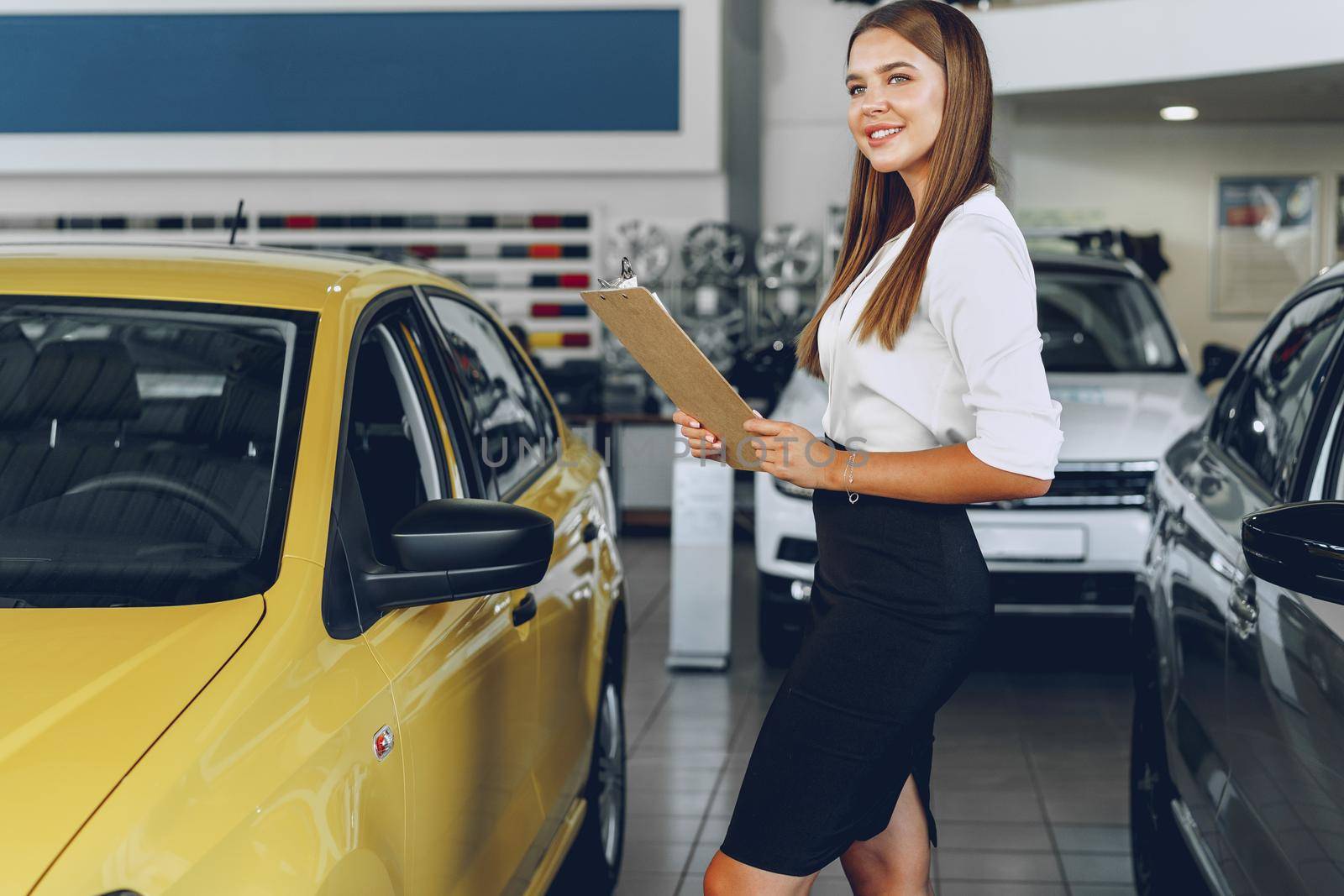 Attractive young female car dealer standing in showroom near a new car