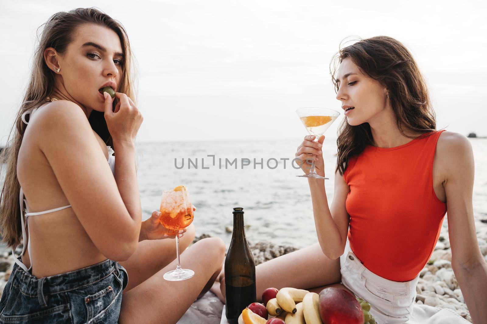 Two young female friends having a picnic on a beach drinking cocktails