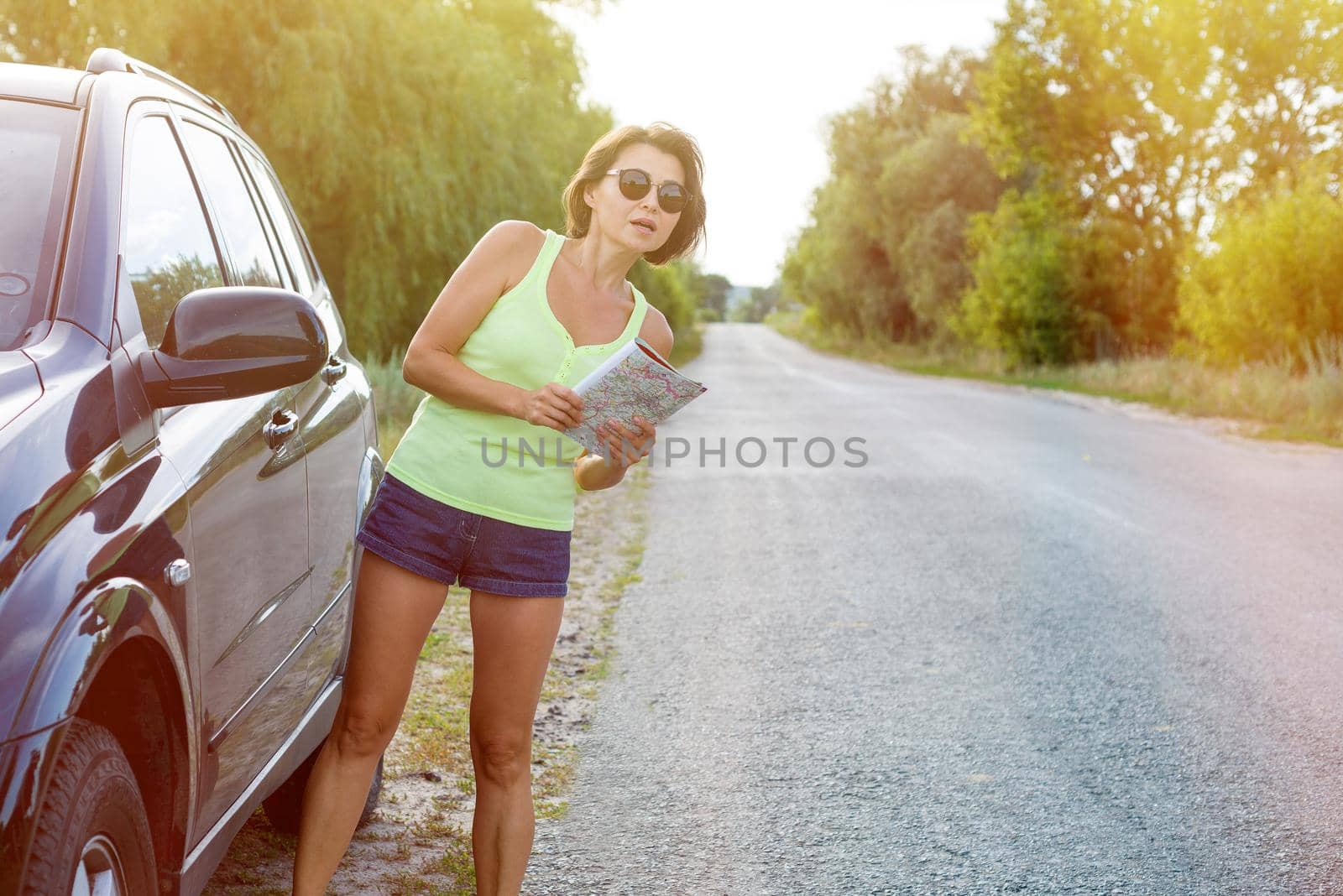 Smiling beautiful woman traveler looking into the tourist map standing near the car in the country road. Travel concept.