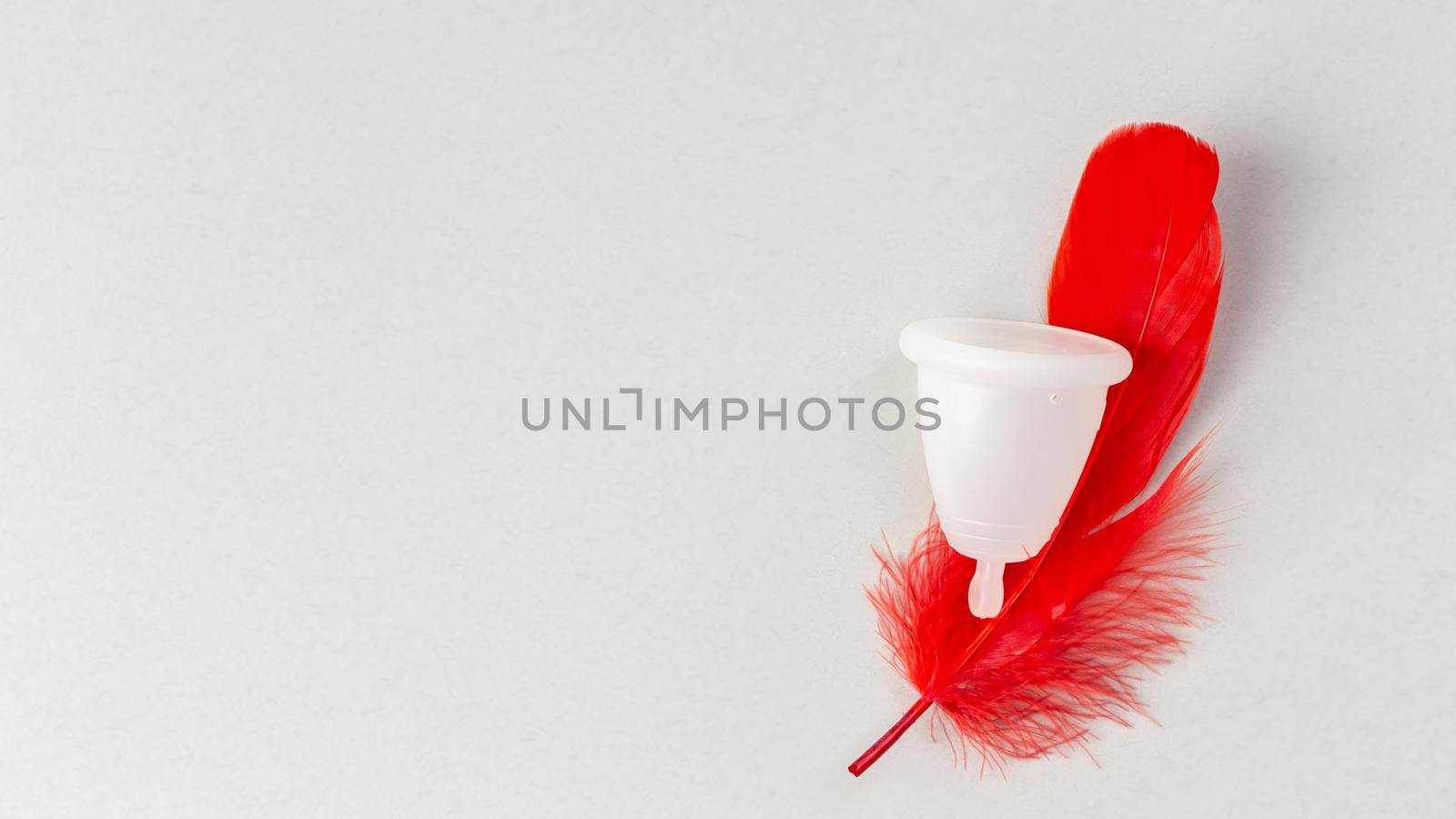 Menstrual cup with red feather over grey background. Periods hygiene product, zero waste alternatives