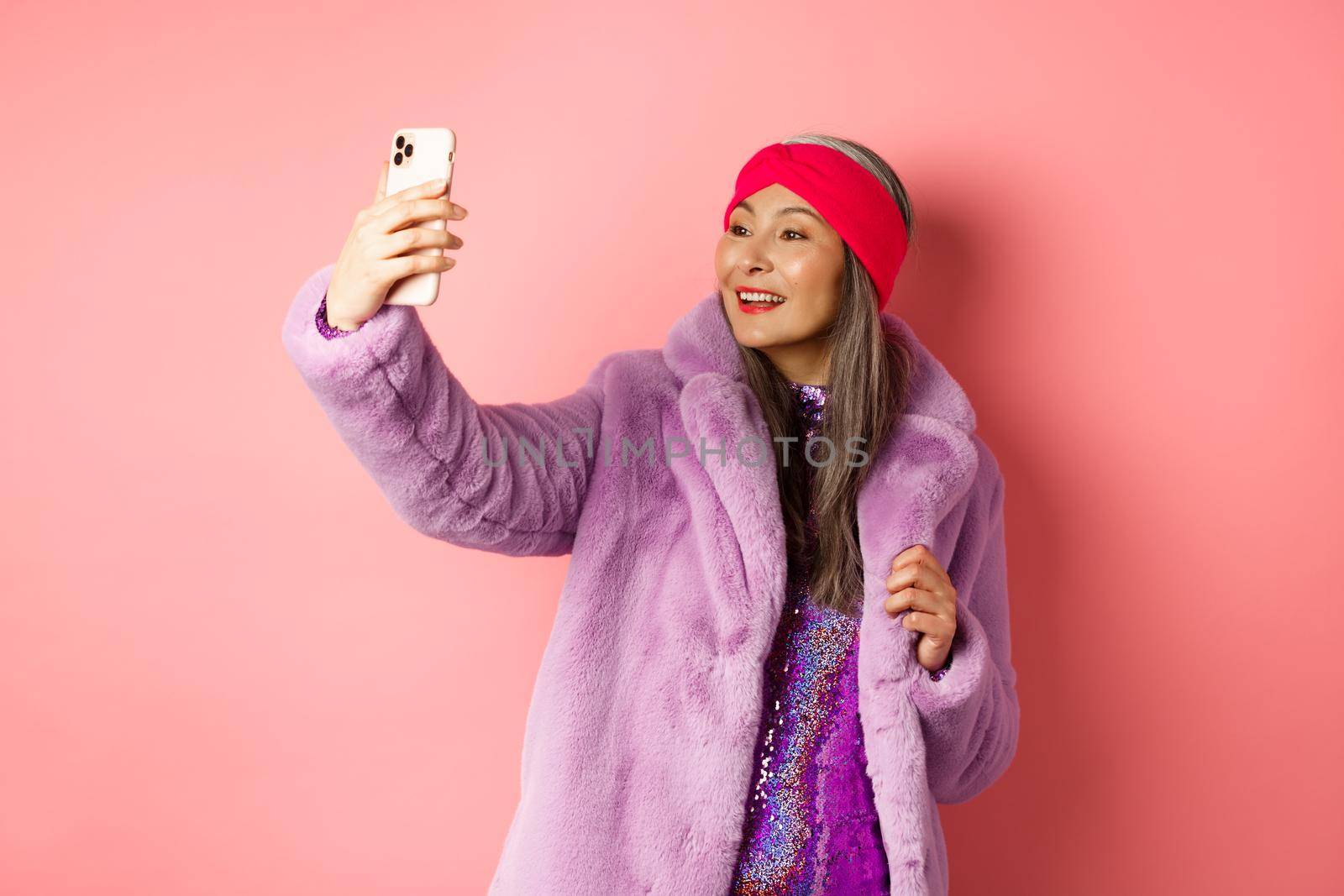Fashion concept. Beautiful asian senior lady taking selfie on smartphone, posing in party glittering dress and purple faux coat, standing over pink background.