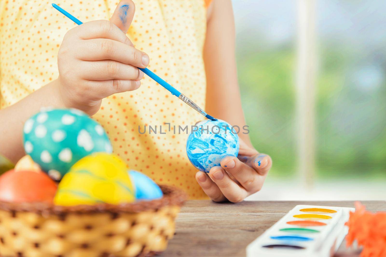 Child is painting egg in blue color for Easter holiday, face is not visible