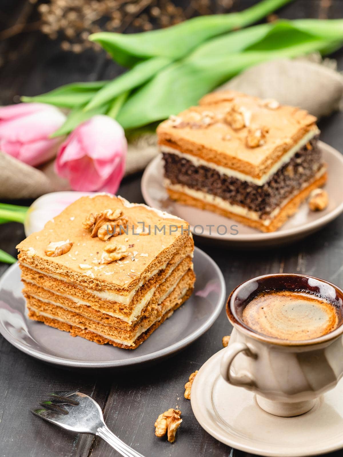 Piece of homemade poppy seed cake and a slice of layered honey cake on the wooden table. Festive set with pastry desserts, cup of coffee and tulip flowers on the background