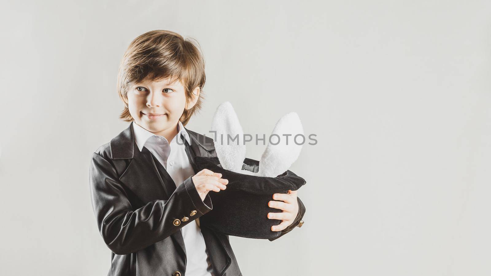 Young magician hiding a toy bunny in his top hat over grey background. Magical performance concept