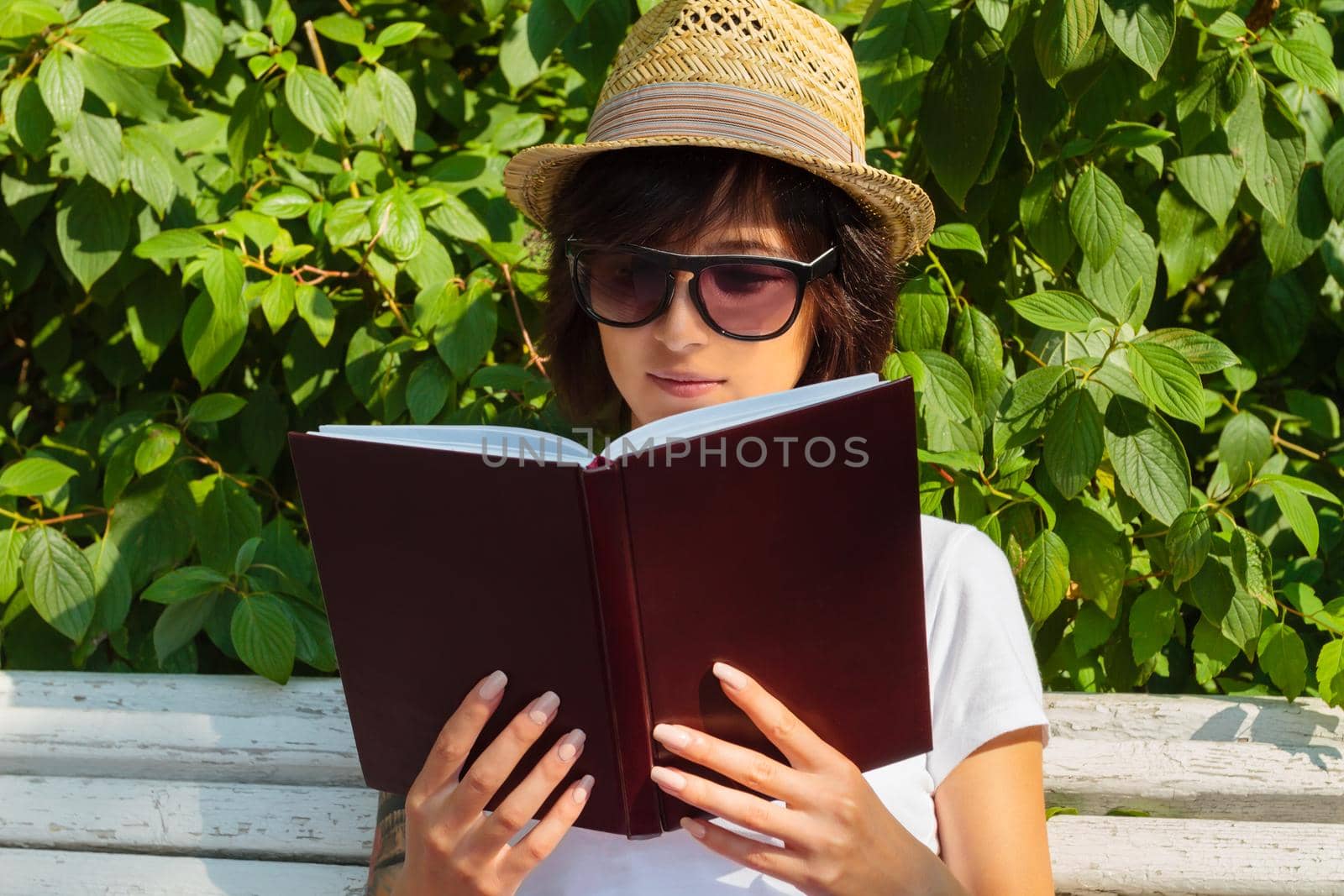 Hipster girl reading a book on a bench in a summer park, front view