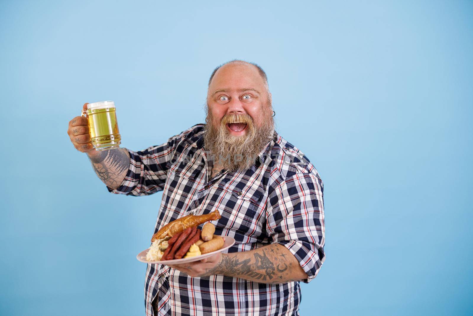 Happy excited bearded man with overweight holds plate of different delicious food and glass of beer standing on light blue background in studio