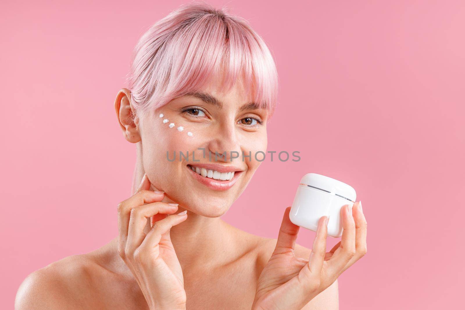 Portrait of gorgeous woman with pink hair and face cream applied on her skin like dots smiling and holding cream in white jar isolated over pink background. Beauty, spa, skin care concept