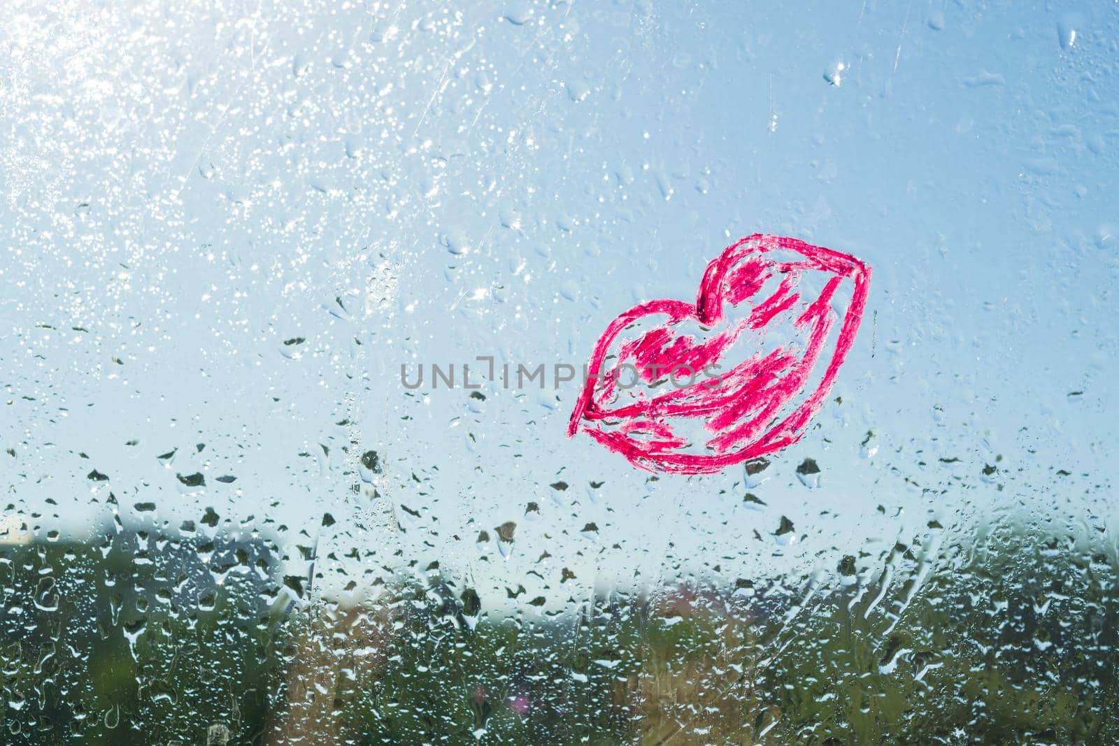Red kiss lips painted with lipstick on the window with water drops. Background blue sunny sky, drops shine in the sun