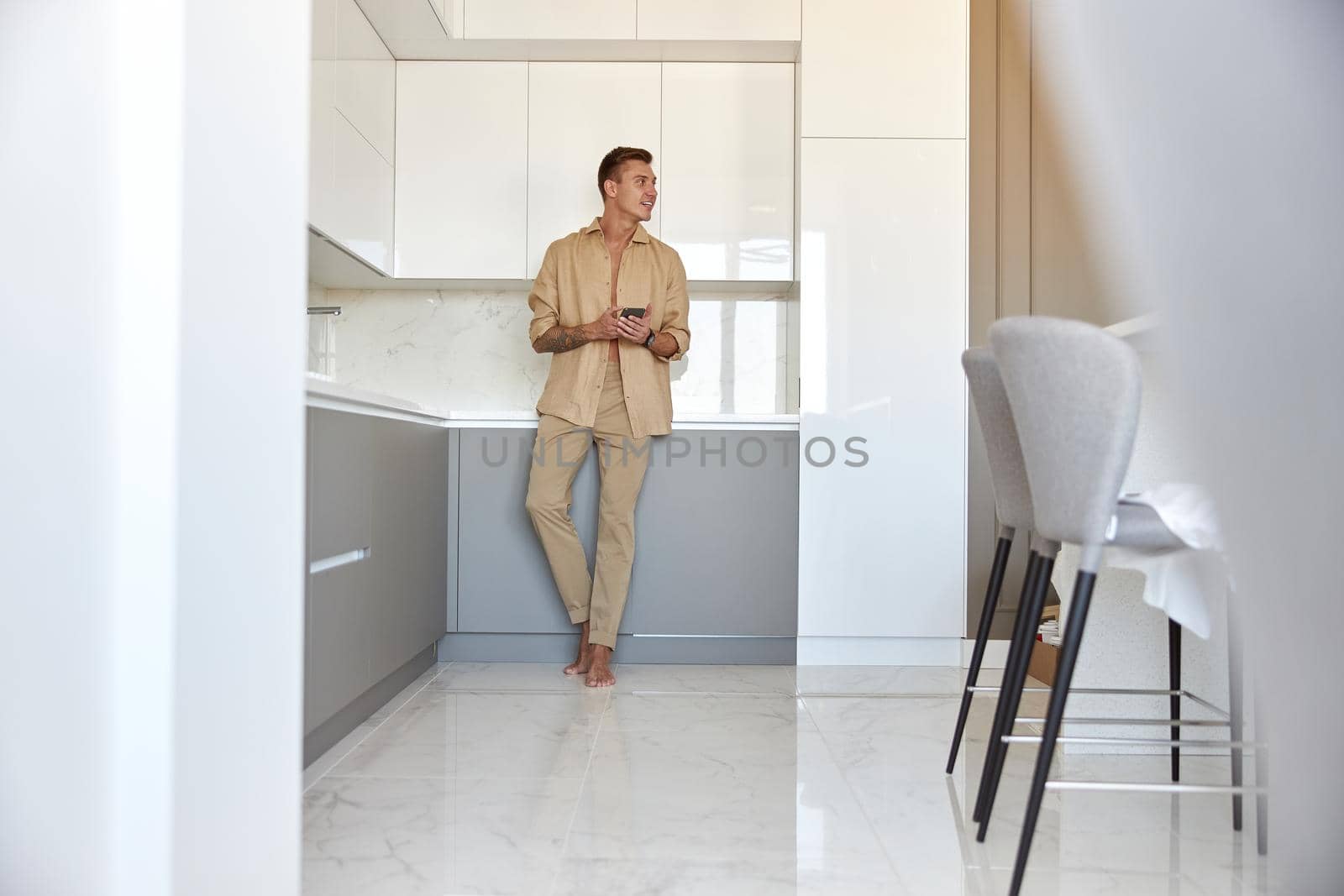 Hadsome smiling man is chilling on the minimalistic kitchen with smartphone