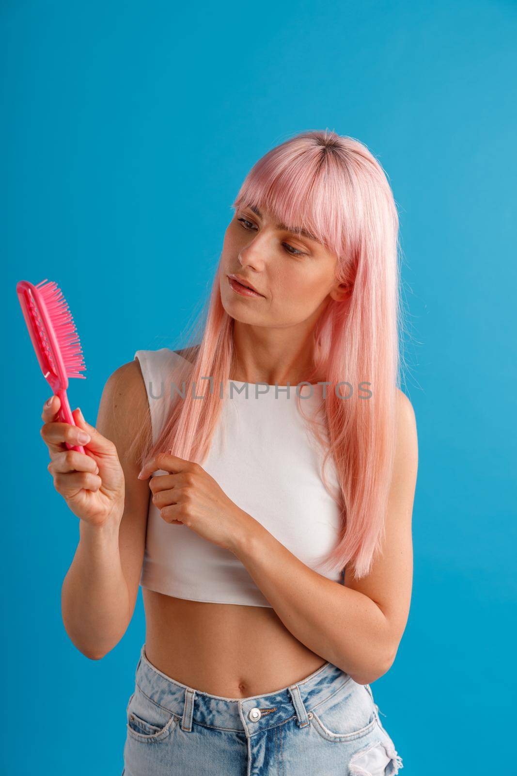 Curious woman with smooth natural long pink hair looking at hair comb while standing isolated over blue studio background by Yaroslav_astakhov