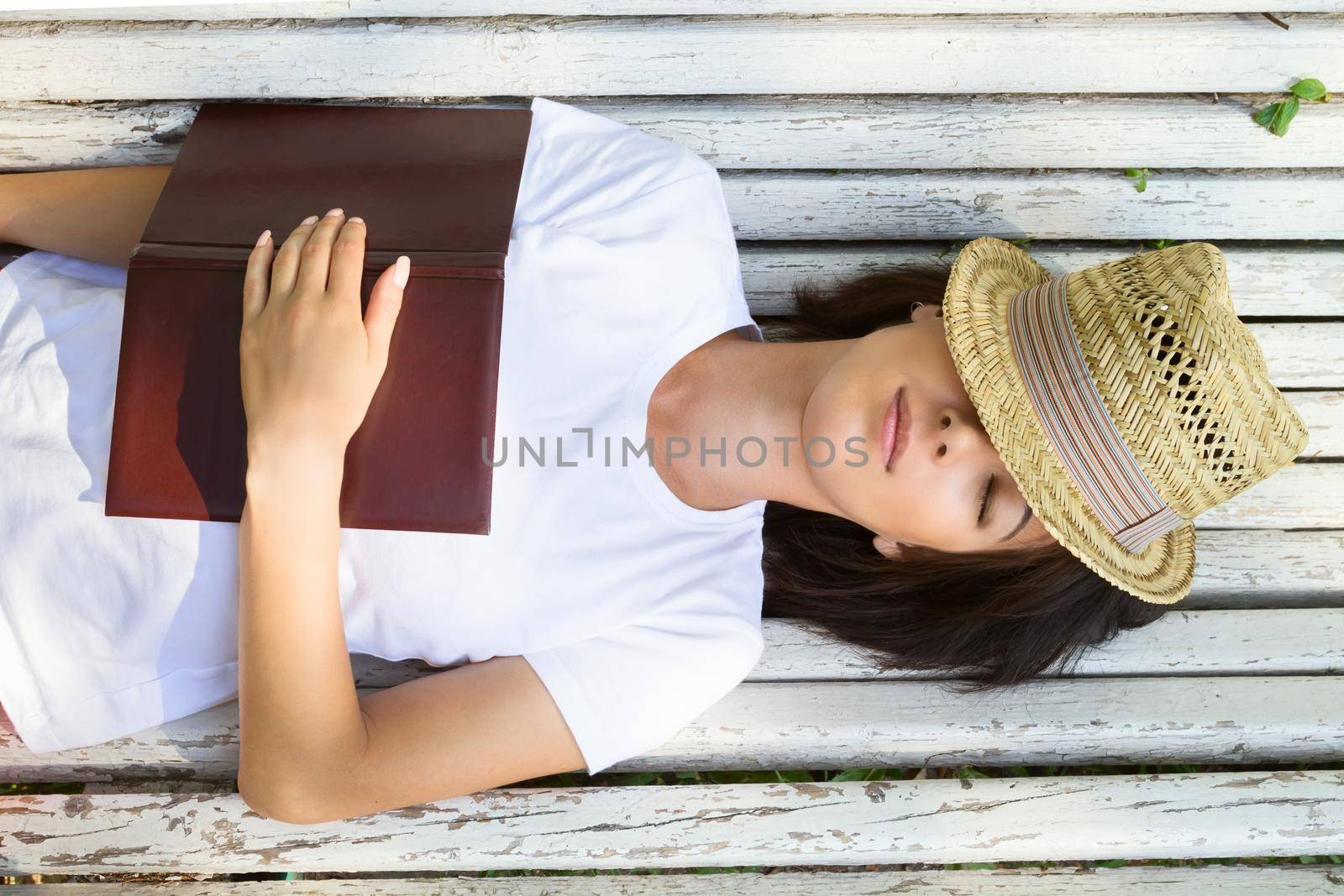 Young woman lies on a bench with a book covered with a hat, top view