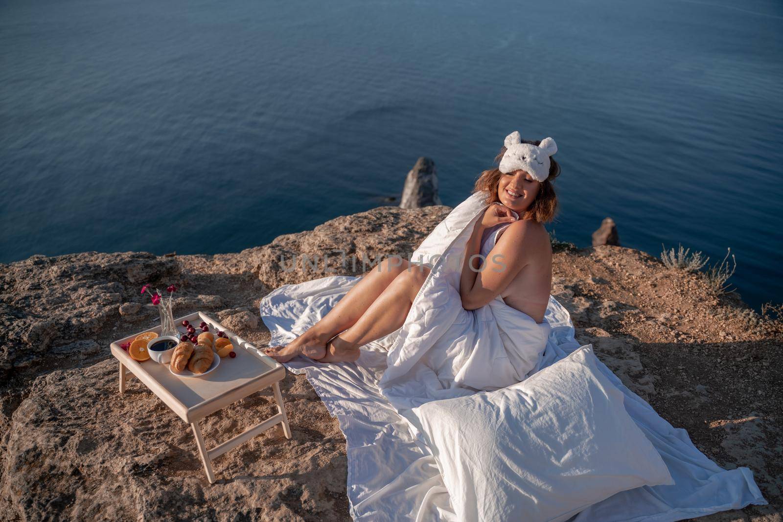 Young woman in sleep mask posing on the seashore. She is wrapped in a white blanket. The coast of the sea at a height, around the sea and rocks, dawn