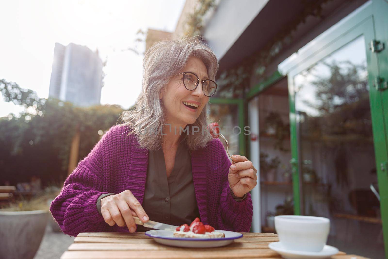 Joyful hoary haired senior lady in purple jacket eats delicious dessert with strawberries sitting at table on outdoors cafe terrace on autumn day