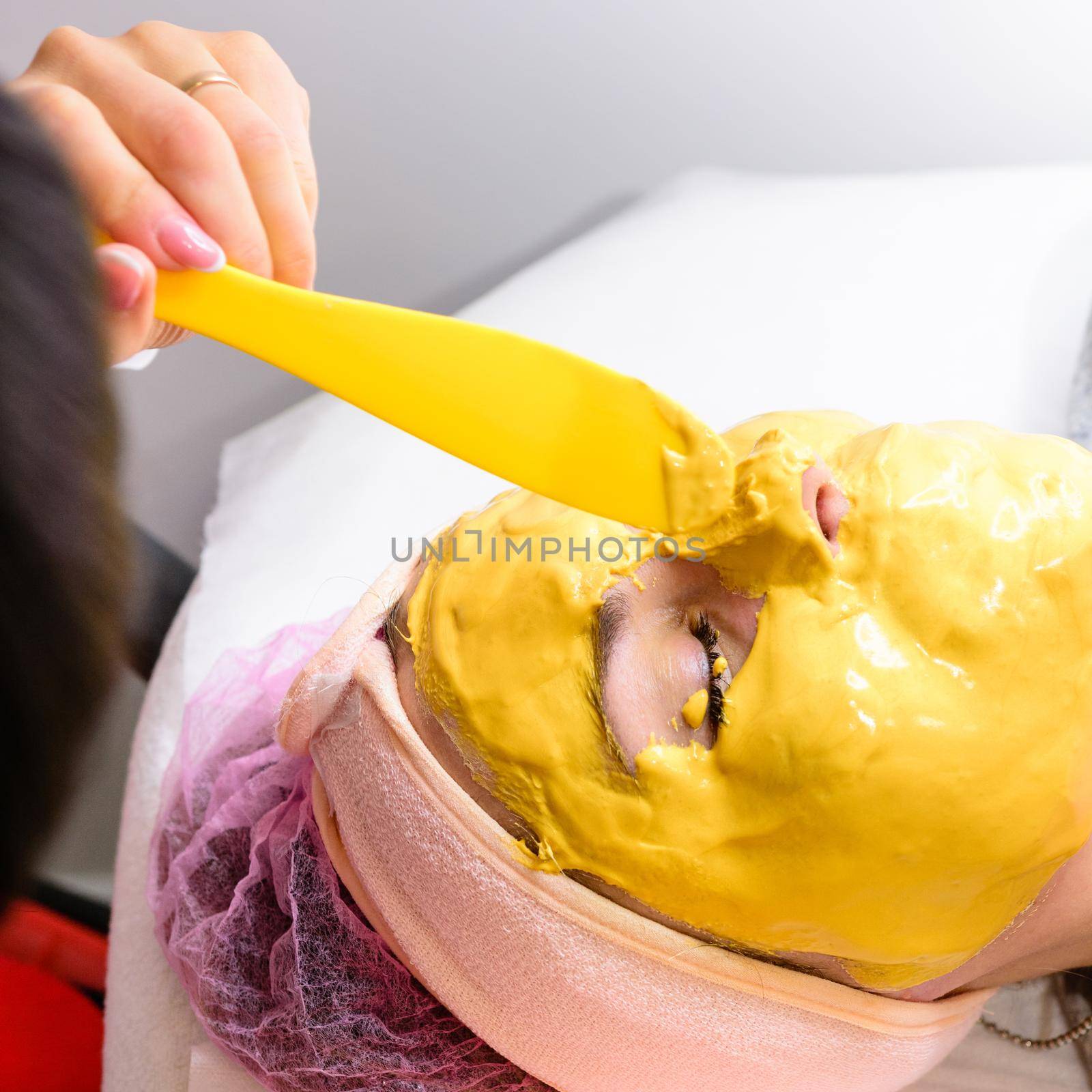 spa treatments in a beauty salon, hand of a professional beautician applying a gold mask on the client's face. by Niko_Cingaryuk
