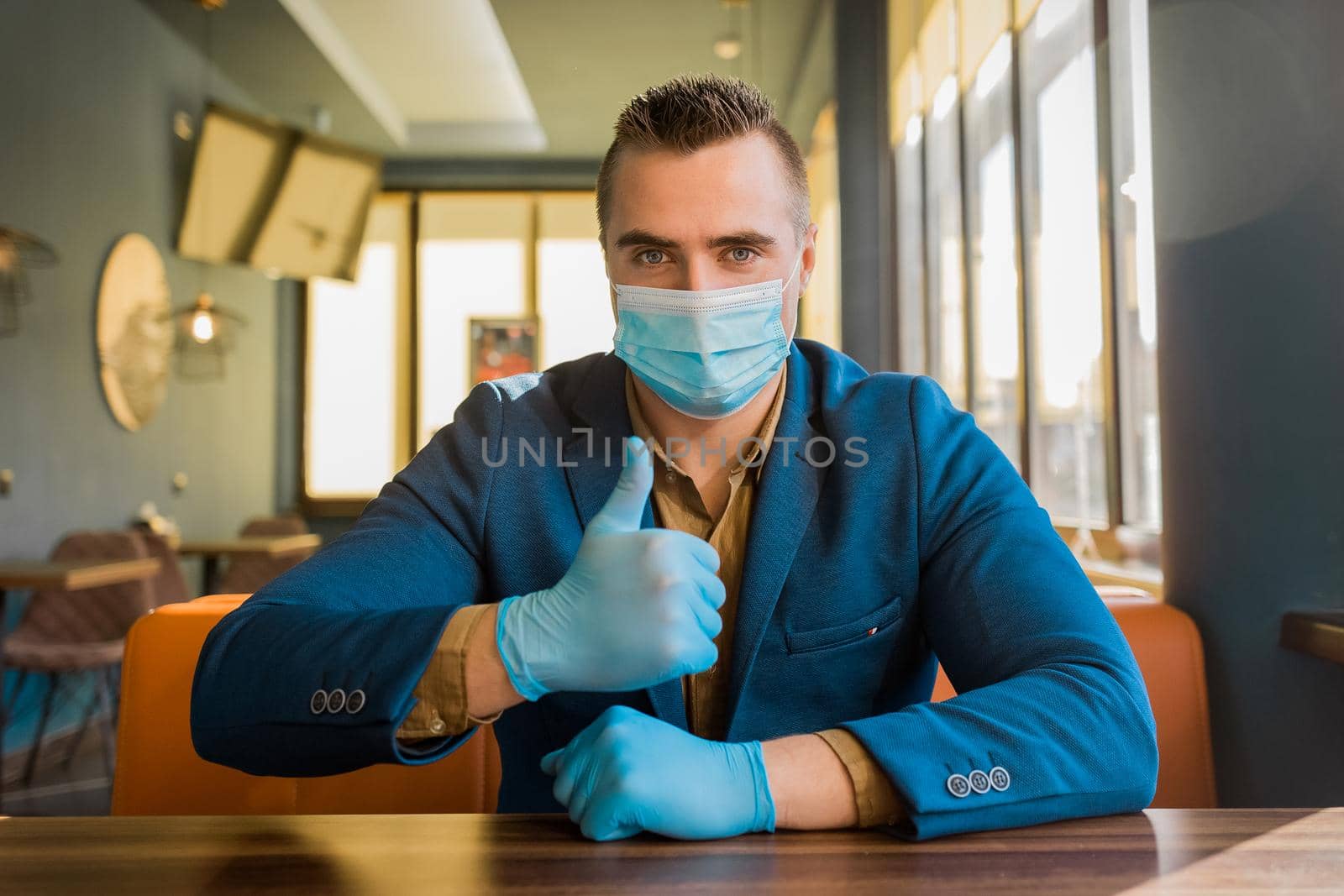 Young caucasian guy businessman in a blue suit and protective gloves and a medical mask shows a cool thumb up.