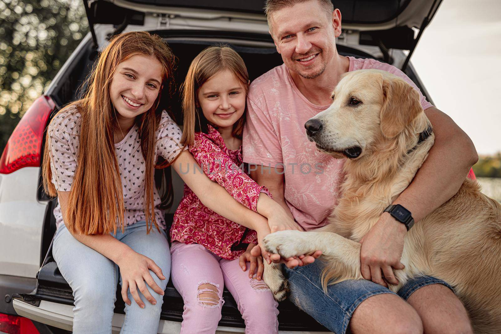 Father with daughters and golden retriever sitting in car trunk on nature