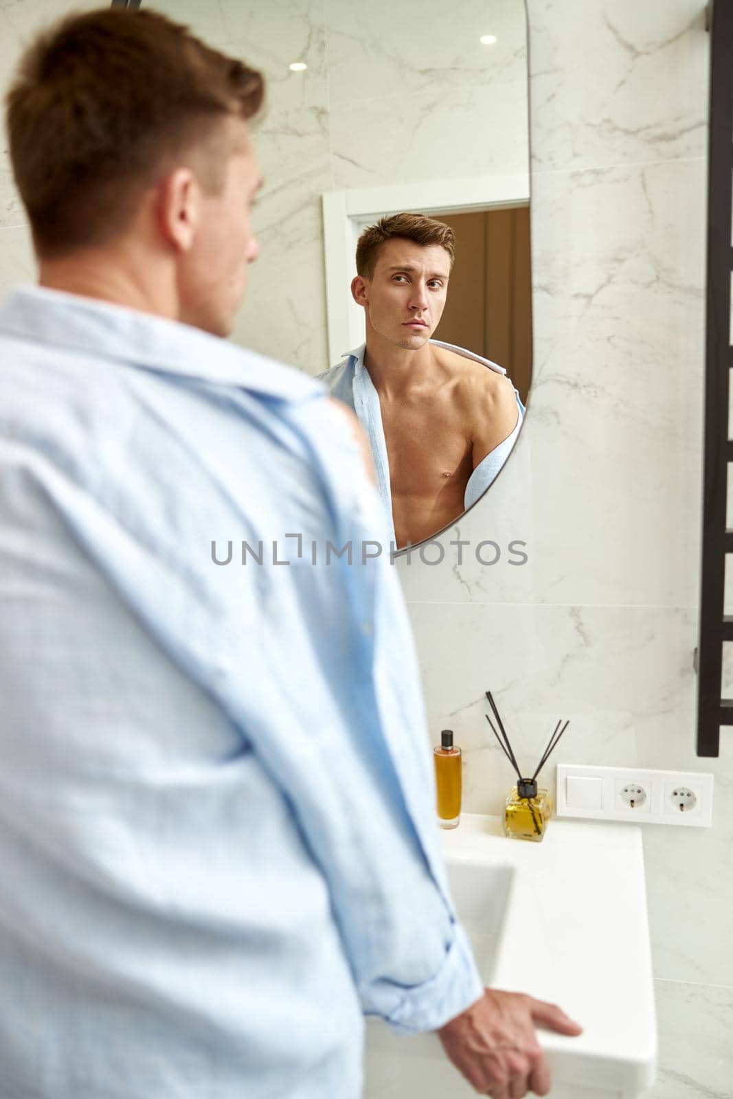 Photo of adult caucasian beautiful man in front of mirror in light modern bathroom