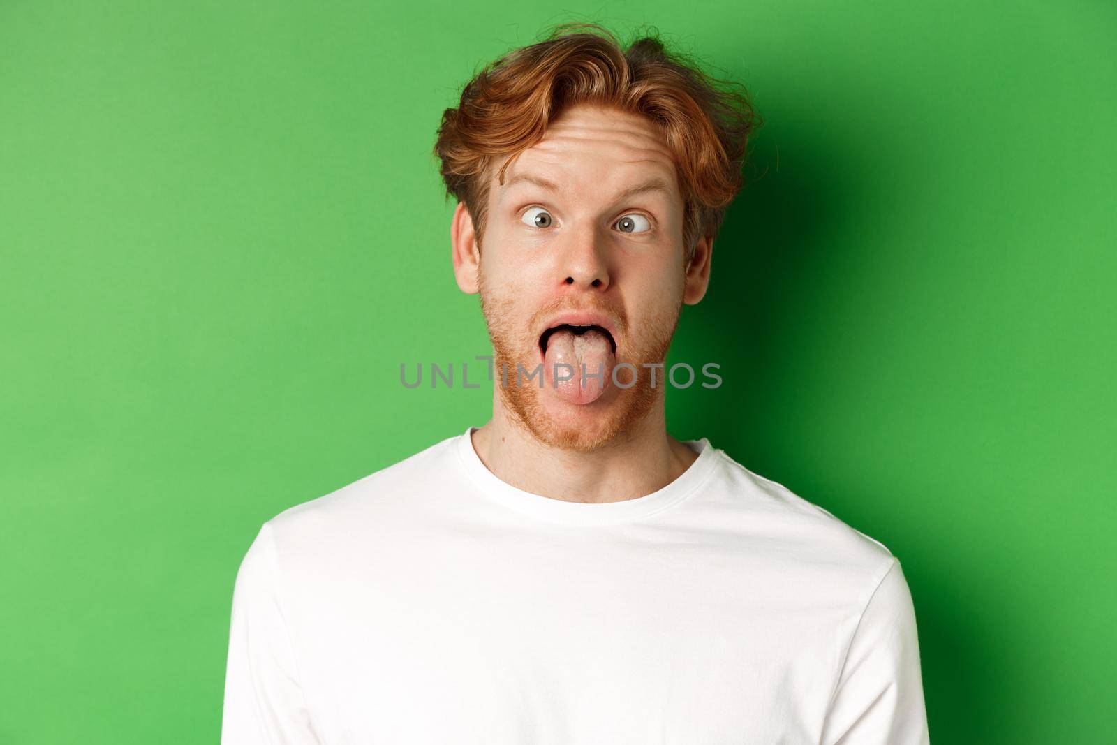 Emotions and fashion concept. Close up of funny redhead man showing silly faces, sticking tongue and squinting, standing over green background by Benzoix