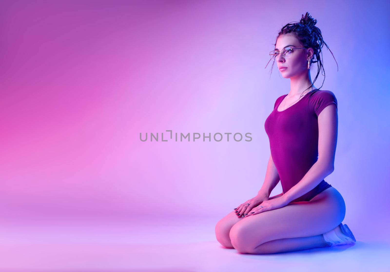 the girl dressed in a bodysuit with dreadlocks on her head is sitting on the floor on a white background in neon light