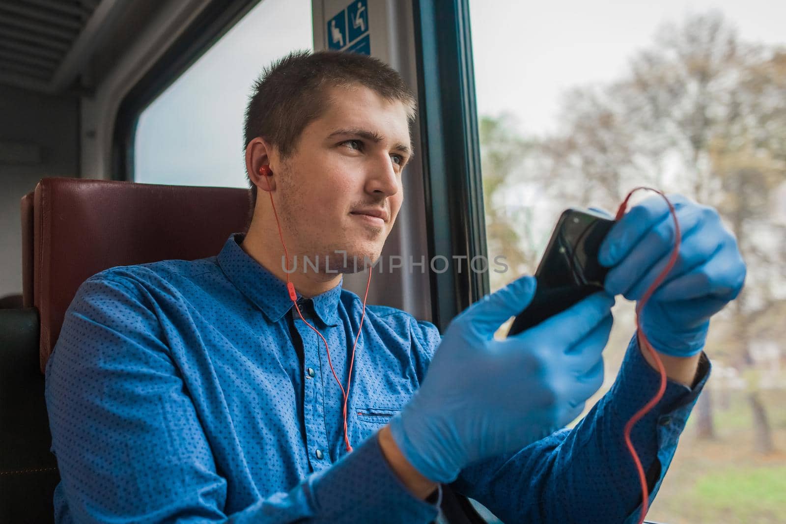 Young european guy in protective medical gloves holds a phone in his hands and listens to music with headphones in a modern electric train by AYDO8