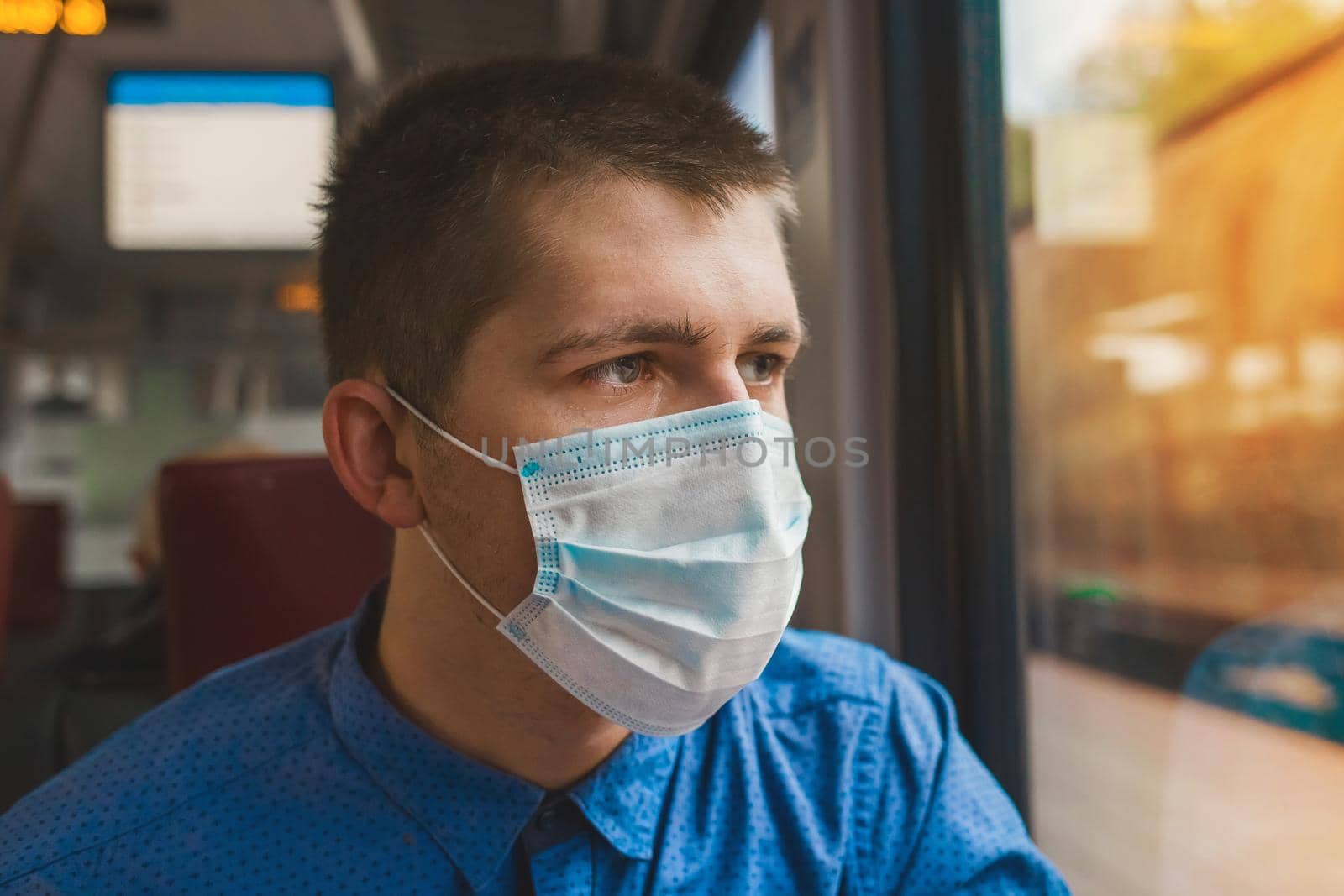 Young Caucasian guy in a protective medical mask looks out the window of a modern electric train at the station.