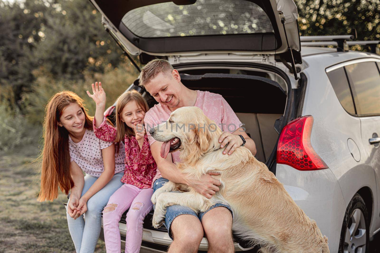 Father with daughters and dog sitting in open car trunk outdoors