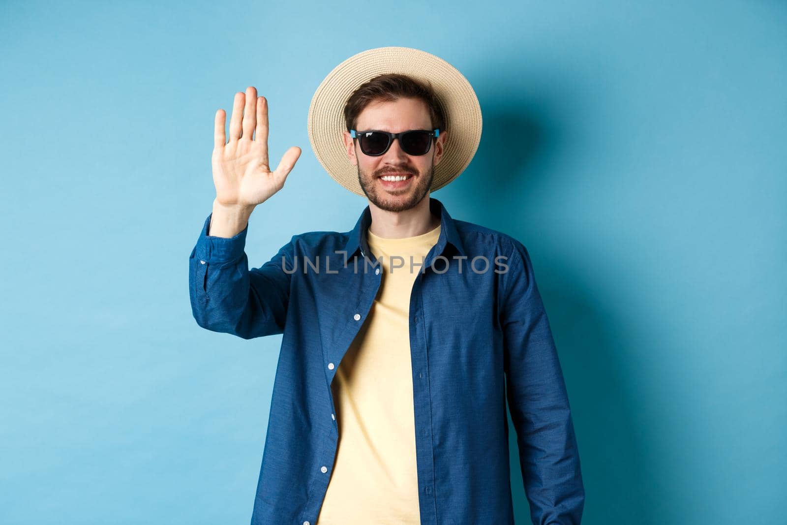 Handsome hipster on summer holiday, waiving hand and smiling, saying hello, wearing sunglasses and straw hat, blue background.