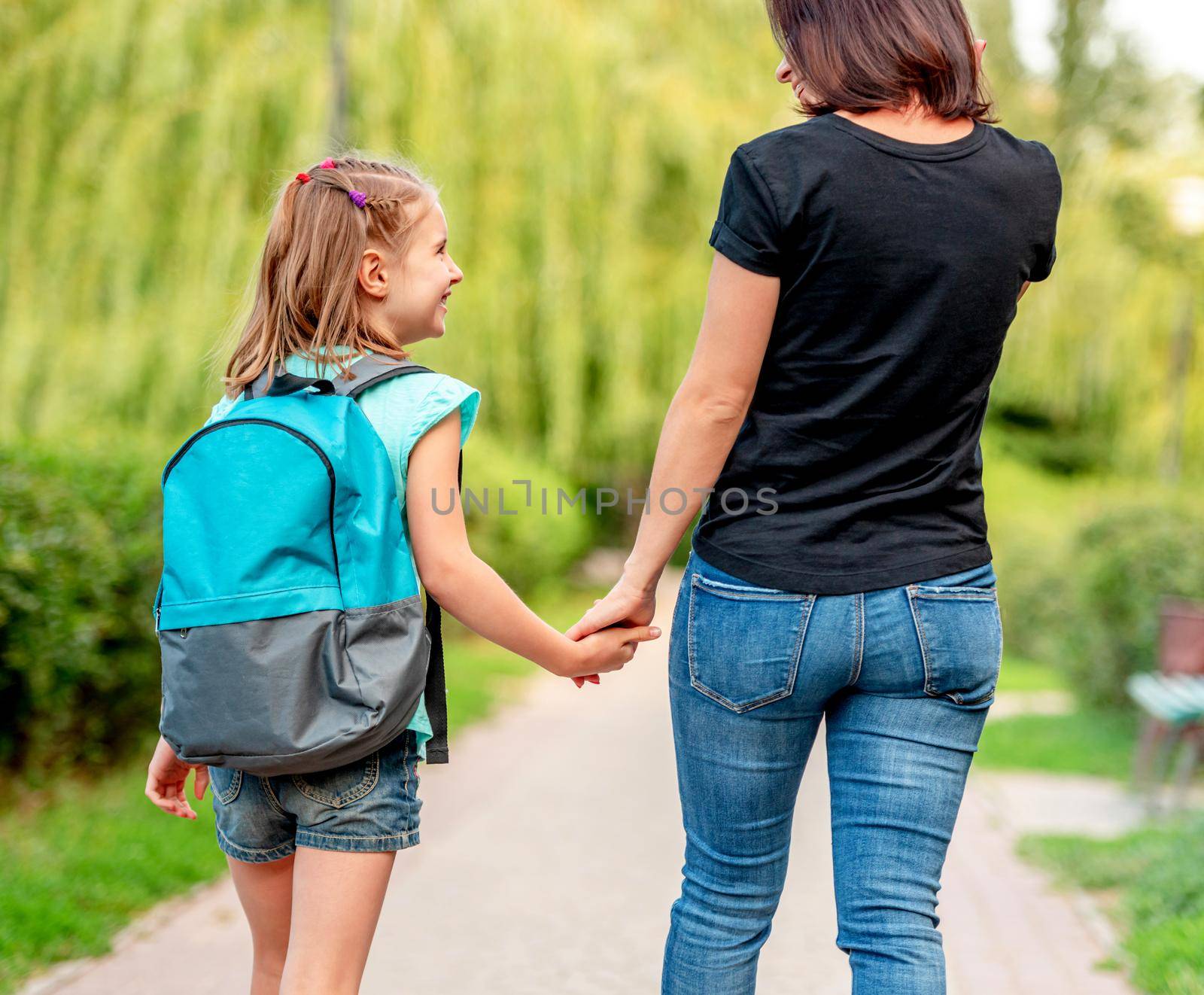 Schoolgirl going home with mother along park alley, rear view