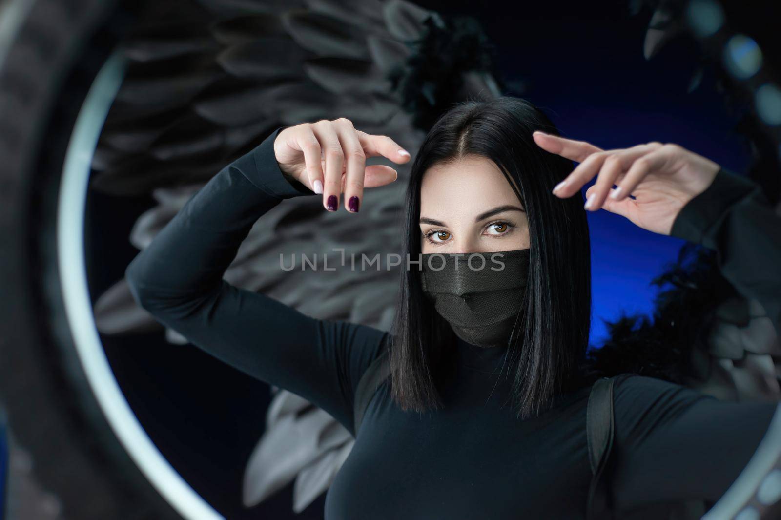 a brunette woman on a background of black wings in a protective mask against coronavirus with hands portrait by Rotozey