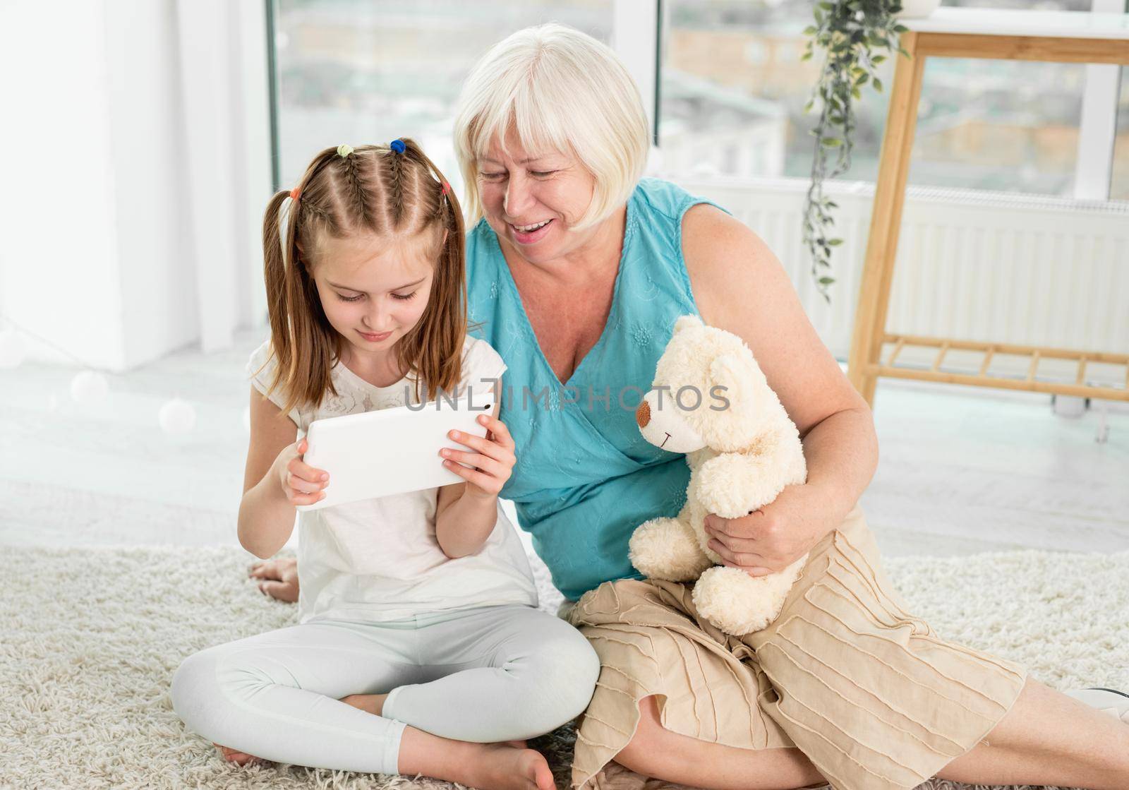 Happy grandmother with little granddaughter using tablet siting on floor in children's room