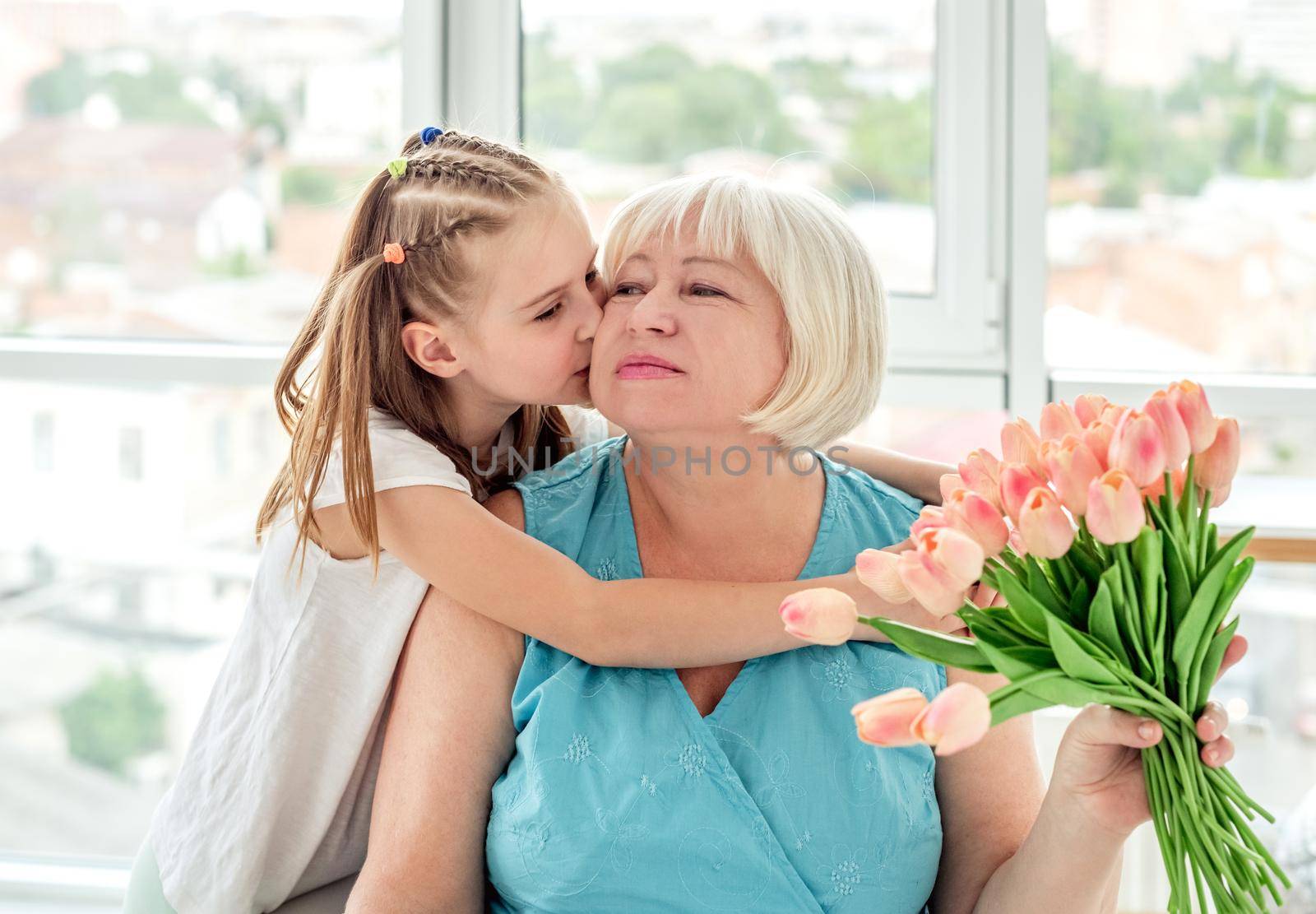 Pretty little girl kissing happy grandmother with bouquet of tulips indoors