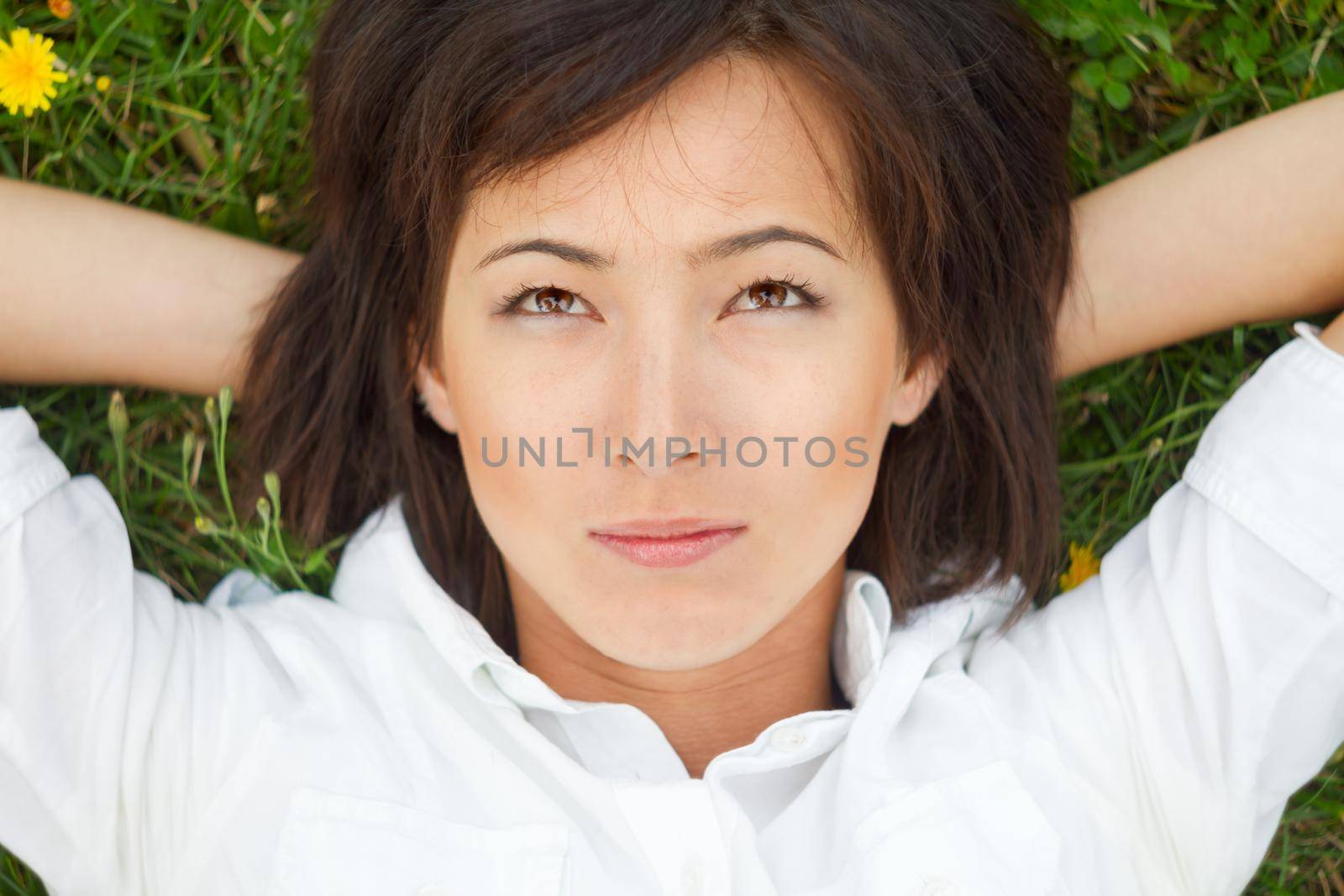 Dreamy young woman lying down on green grass with hands behind her head.