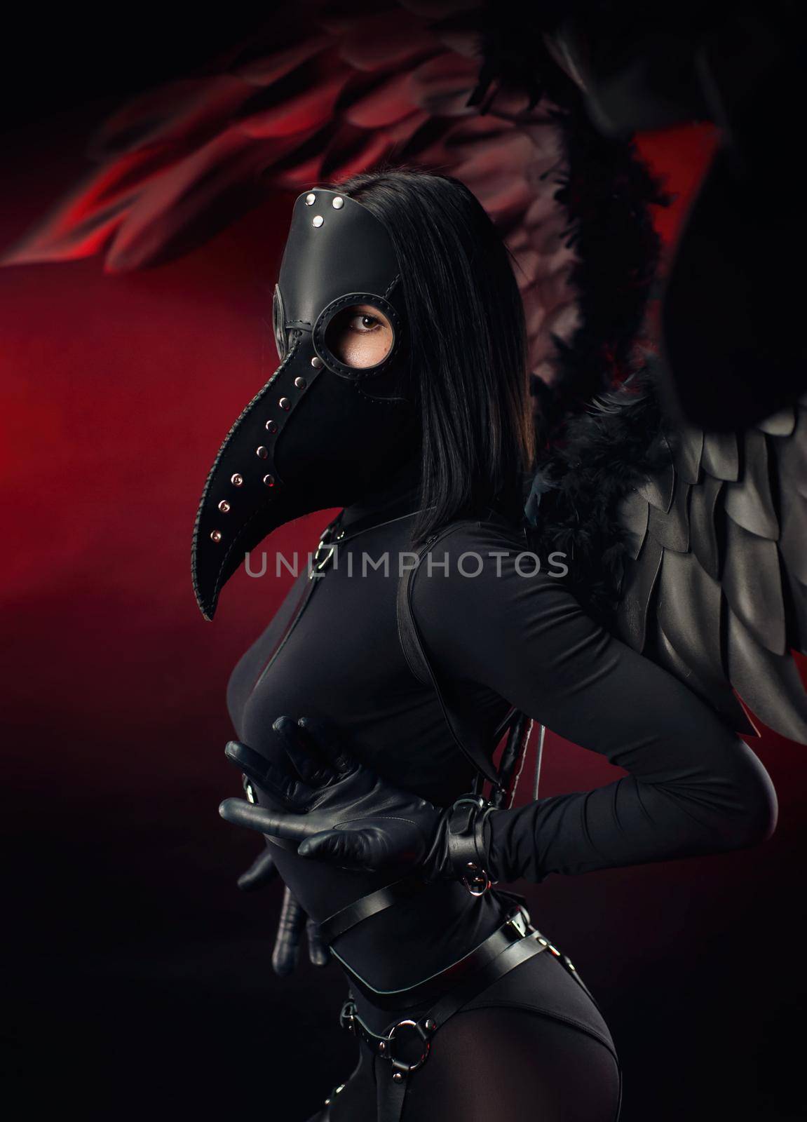 the woman wearing a plague doctor mask with leather straps and black wings on a dark red background