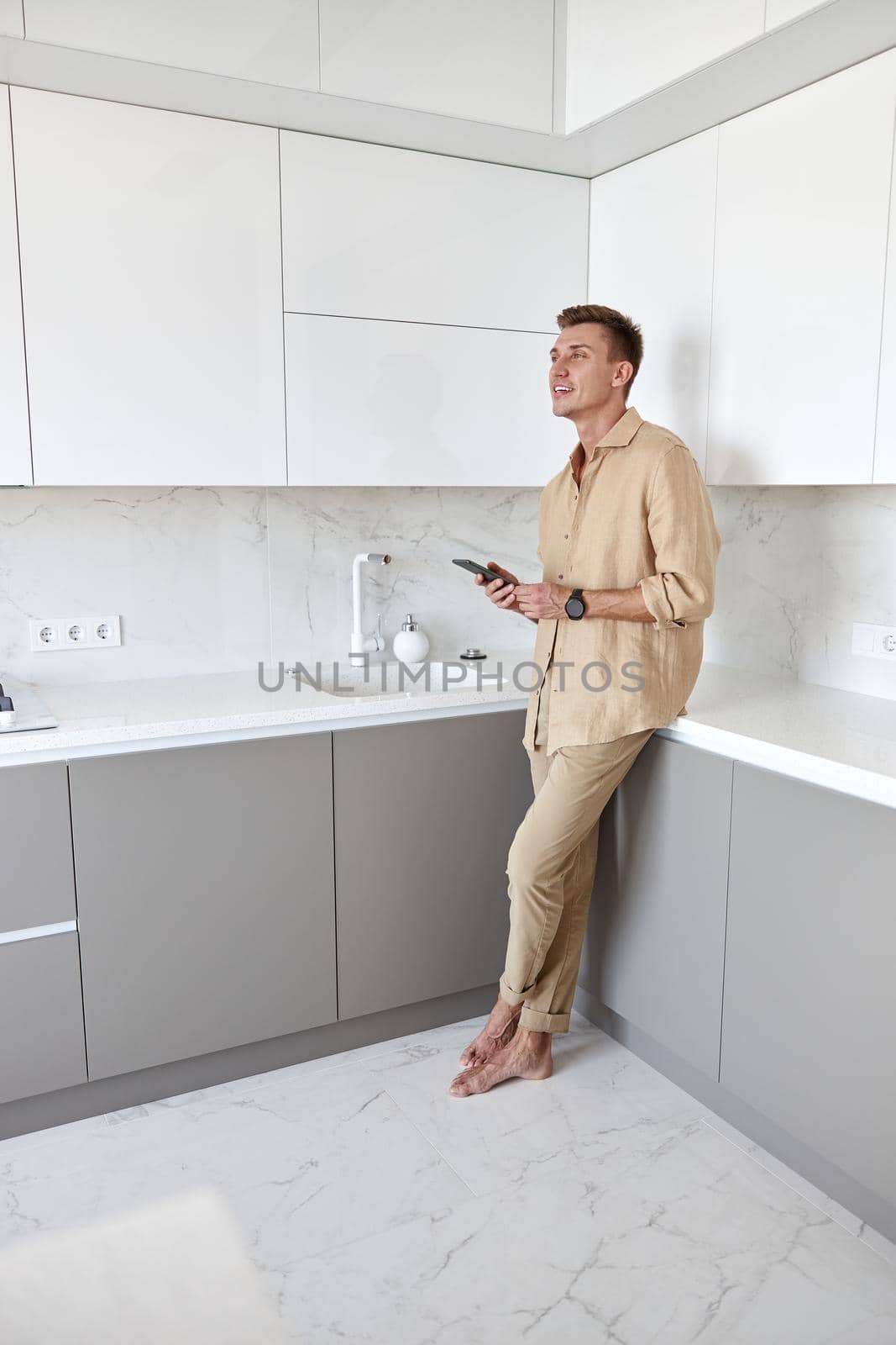 Hadsome smiling man is chilling on the minimalistic kitchen with smartphone by Yaroslav_astakhov